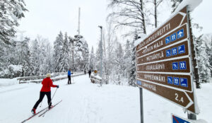 Hiihtäjiä lumisessa maisemassa, reittikyltin vieressä.
