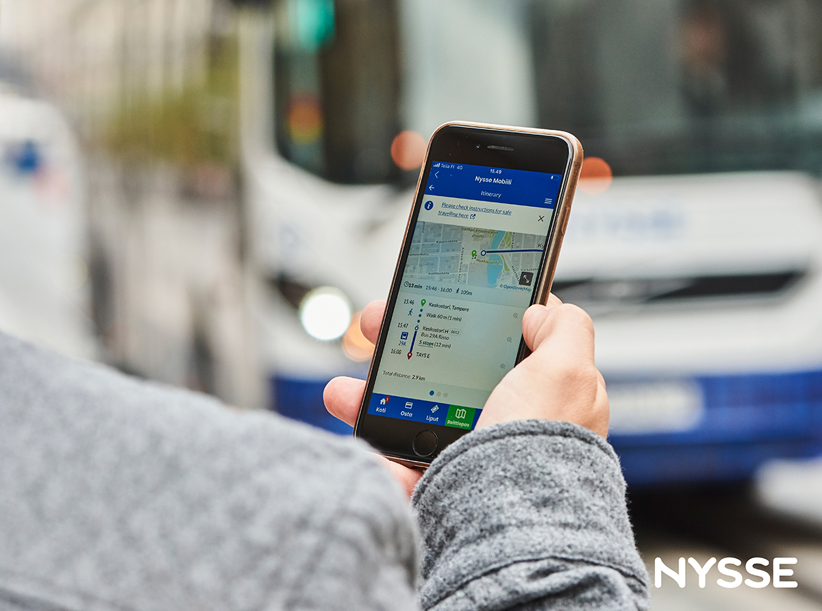 Henkilö katsoo kännykästä Nysse-reittiopasta, linja-auto saapuu pysäkille.